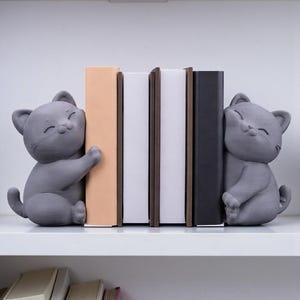 May include: Gray cat bookends holding books on a white shelf. The cat bookends are smiling and hugging the books. The books are various colors, including beige, white, and black. The shelf is white.