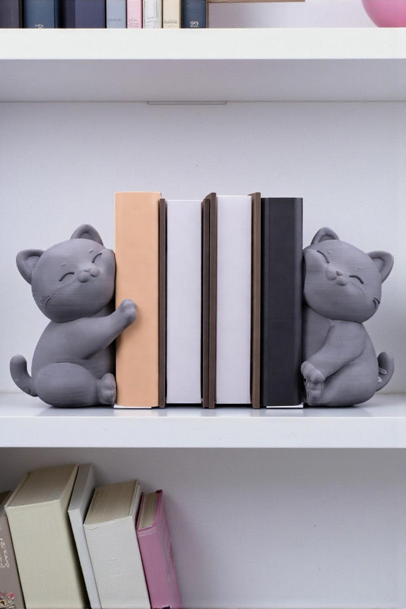 May include: Gray cat bookends holding books on a white shelf. The cat bookends are smiling and hugging the books. The books are various colors, including beige, white, and black. The shelf is white.