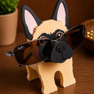 May include: A whimsical dog-shaped eyeglass holder in tan, black, and pink. The holder features a pug-like design with sunglasses resting on its nose. The sunglasses have a silver frame and brown lenses. The holder is made of foam.