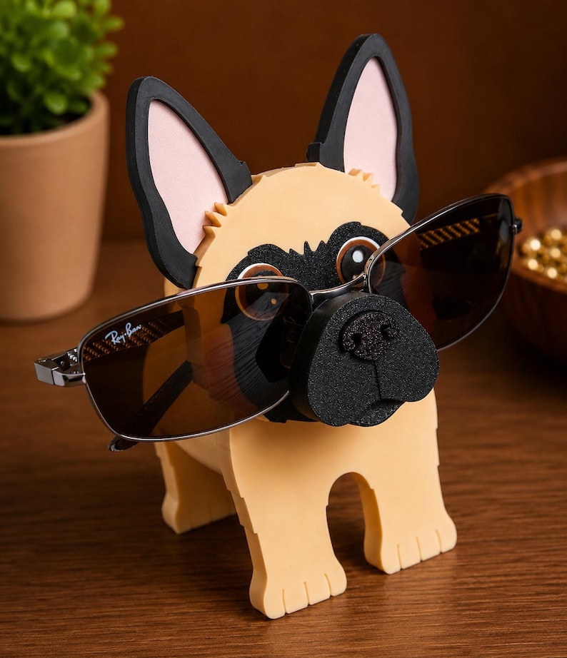 May include: A whimsical dog-shaped eyeglass holder in tan, black, and pink. The holder features a pug-like design with sunglasses resting on its nose. The sunglasses have a silver frame and brown lenses. The holder is made of foam.