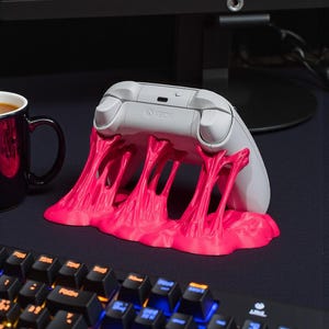 May include: A white Xbox controller rests on a vibrant pink, melted-looking stand. The stand has a unique, dripping design. A black keyboard and a mug of coffee are in the background.