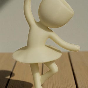 May include: A cream-colored figurine of a ballerina in a graceful pose. The figure has a rounded head, arms outstretched, and a tutu. The figurine is standing on a round base. The background is a wooden surface.