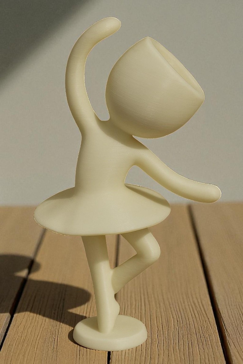 May include: A cream-colored figurine of a ballerina in a graceful pose. The figure has a rounded head, arms outstretched, and a tutu. The figurine is standing on a round base. The background is a wooden surface.