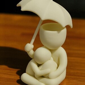 May include: A cream-colored figurine depicting a person holding an umbrella over another person in a seated embrace. The umbrella is held aloft, providing shelter. The sculpture is smooth and minimalist, with rounded forms.
