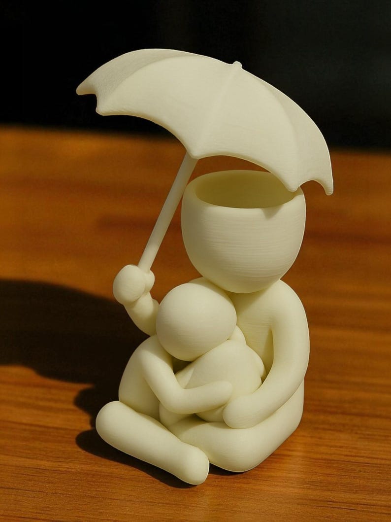 May include: A cream-colored figurine depicting a person holding an umbrella over another person in a seated embrace. The umbrella is held aloft, providing shelter. The sculpture is smooth and minimalist, with rounded forms.