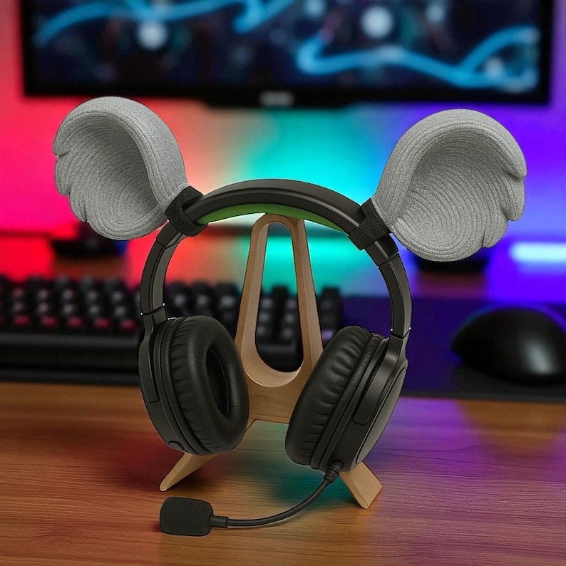May include: Black over-ear headphones with a boom microphone, resting on a wooden stand. The headphones have gray ear covers and are positioned on a wooden stand. The background features a keyboard, mouse, and a monitor displaying colorful lights.