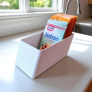 May include: A white, angled container holding several packets of Dr. Oetker Natron baking soda. The container sits on a countertop near a window and a kitchen faucet. The packets are orange and blue.