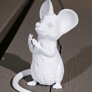 May include: A white mouse figurine with large ears and a long tail. The mouse is in a standing position with its paws raised. The figurine is made of a smooth material and sits on a wooden surface.