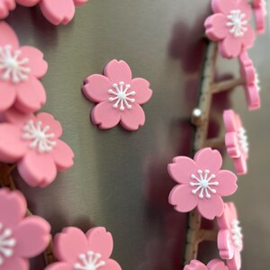 May include: Pink cherry blossom magnets with white centers are displayed on a metallic surface. The magnets are arranged in clusters and along a branch-like structure. The image showcases a close-up view of the decorative floral magnets.