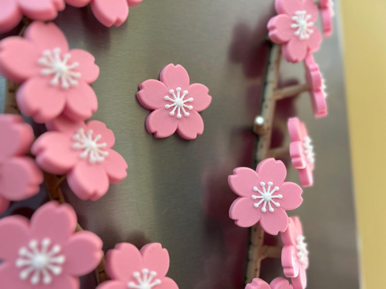 May include: Pink cherry blossom magnets with white centers are displayed on a metallic surface. The magnets are arranged in clusters and along a branch-like structure. The image showcases a close-up view of the decorative floral magnets.