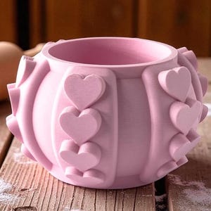 May include: A light pink planter with heart-shaped detailing. The round pot has a textured surface and is made of a solid material. The hearts are evenly spaced around the exterior, adding a decorative touch. The planter sits on a wooden surface.