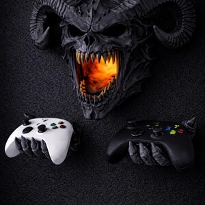 Puede incluir: Decoraci&oacute;n de pared de calavera demon&iacute;aca gris oscuro con cuernos grandes y un interior ardiente. Debajo, un mando de juego blanco y negro sostenido por manos esquel&eacute;ticas.