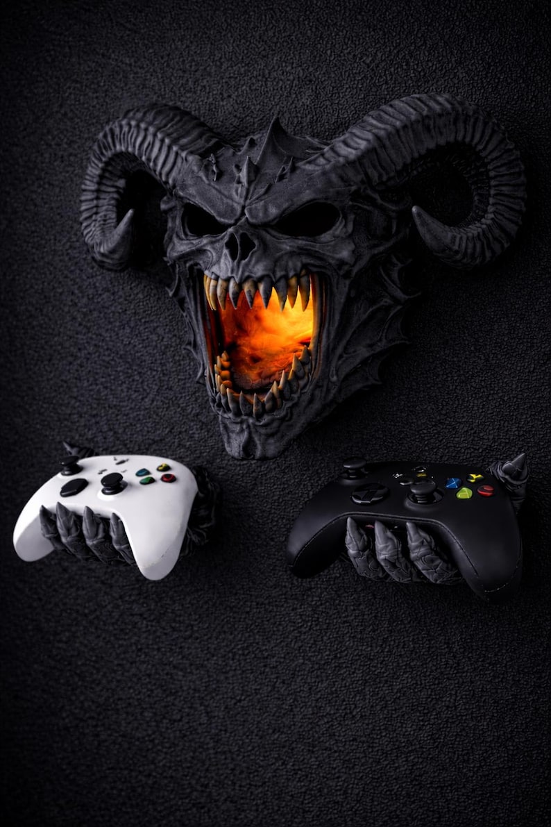 Puede incluir: Decoraci&oacute;n de pared de calavera demon&iacute;aca gris oscuro con cuernos grandes y un interior ardiente. Debajo, un mando de juego blanco y negro sostenido por manos esquel&eacute;ticas.