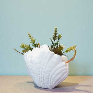 May include: A white seashell-shaped vase filled with greenery and small white flowers. The vase has a textured surface and sits on a beige surface against a light blue background. The arrangement includes sprigs of green and yellow.