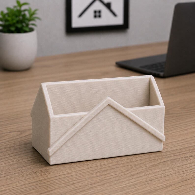 Pu&ograve; includere: Un organizer da scrivania beige a forma di casa. L'organizer ha una base rettangolare e un design a tetto spiovente. &Egrave; realizzato in un materiale strutturato e poggia su una scrivania di legno. Una piccola pianta in vaso e un laptop sono sullo sfondo.