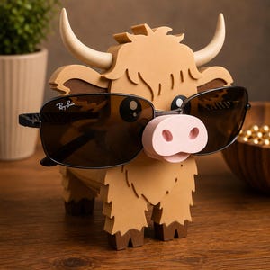May include: A whimsical, brown Highland cow-shaped eyeglass holder with a light pink nose and cream-colored horns. The holder is wearing black Ray-Ban sunglasses. The holder is made of wood and has a shaggy appearance.