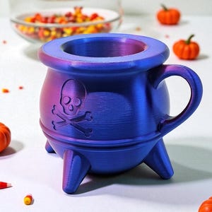 May include: A blue and purple cauldron-shaped mug with a skull and crossbones design. The mug has a handle and three legs. The mug is a Halloween-themed item.