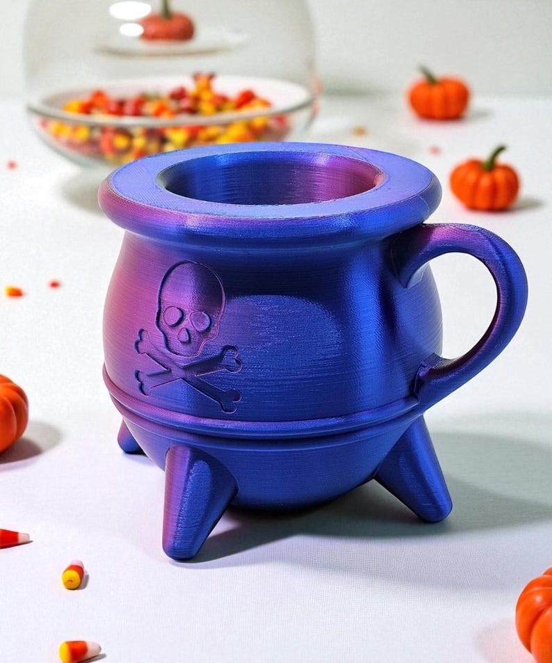 May include: A blue and purple cauldron-shaped mug with a skull and crossbones design. The mug has a handle and three legs. The mug is a Halloween-themed item.