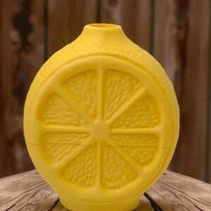 May include: A bright yellow, lemon-shaped decorative bottle. The bottle features a detailed cross-section of a lemon slice on its front. The bottle has a narrow neck and a rounded base. The background is a wooden surface.