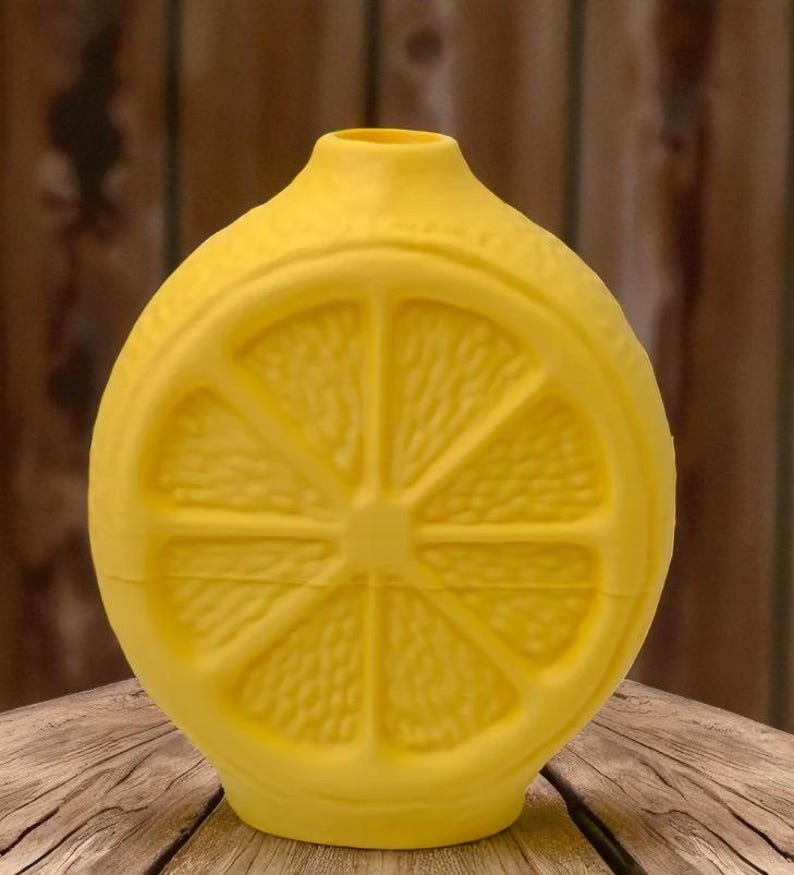 May include: A bright yellow, lemon-shaped decorative bottle. The bottle features a detailed cross-section of a lemon slice on its front. The bottle has a narrow neck and a rounded base. The background is a wooden surface.