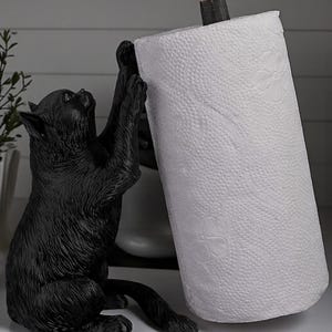 May include: A black cat-shaped paper towel holder. The cat is sculpted in a playful pose, reaching up to hold a roll of white paper towels. A small yellow bird sits atop the holder. A decorative and functional kitchen accessory.