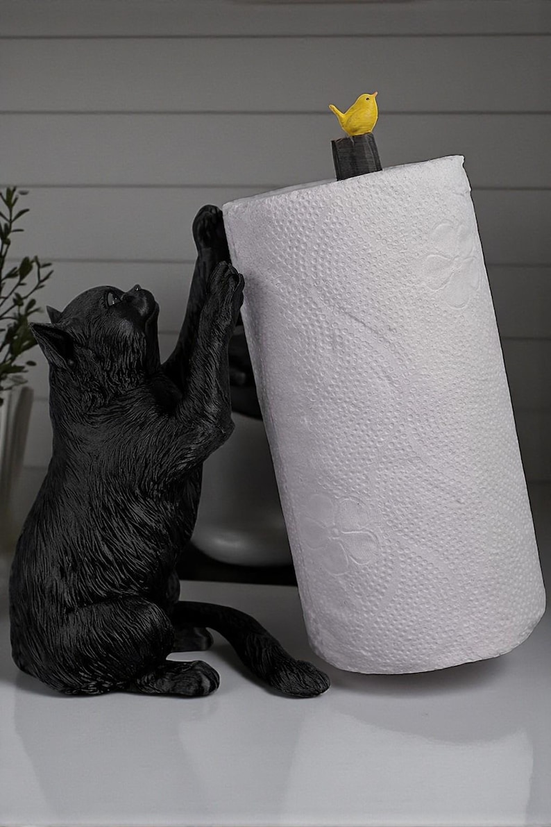 May include: A black cat-shaped paper towel holder. The cat is sculpted in a playful pose, reaching up to hold a roll of white paper towels. A small yellow bird sits atop the holder. A decorative and functional kitchen accessory.
