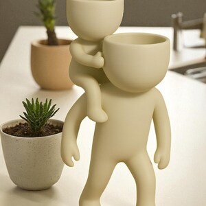 Op de afbeelding: Een cr&egrave;mekleurige decoratieve plantenbak in de vorm van een persoon die een andere persoon op de schouders draagt. De bovenkant van elke figuur is een kleine, open kom om in te planten. Twee kleine potplanten staan op de achtergrond.