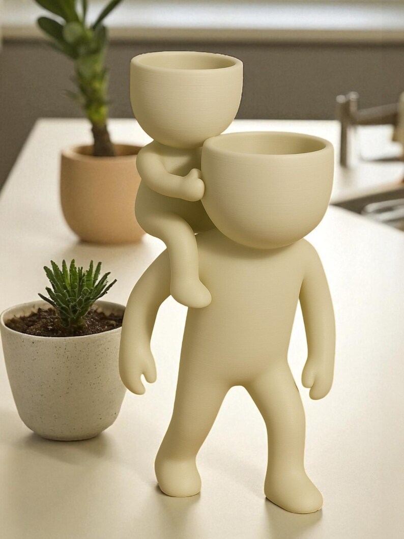 Op de afbeelding: Een cr&egrave;mekleurige decoratieve plantenbak in de vorm van een persoon die een andere persoon op de schouders draagt. De bovenkant van elke figuur is een kleine, open kom om in te planten. Twee kleine potplanten staan op de achtergrond.