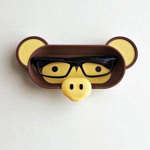 May include: A brown and yellow eyeglasses holder shaped like a monkey face. The holder has a tray for glasses, yellow ears, and a yellow nose. Black eyeglasses rest inside the tray.