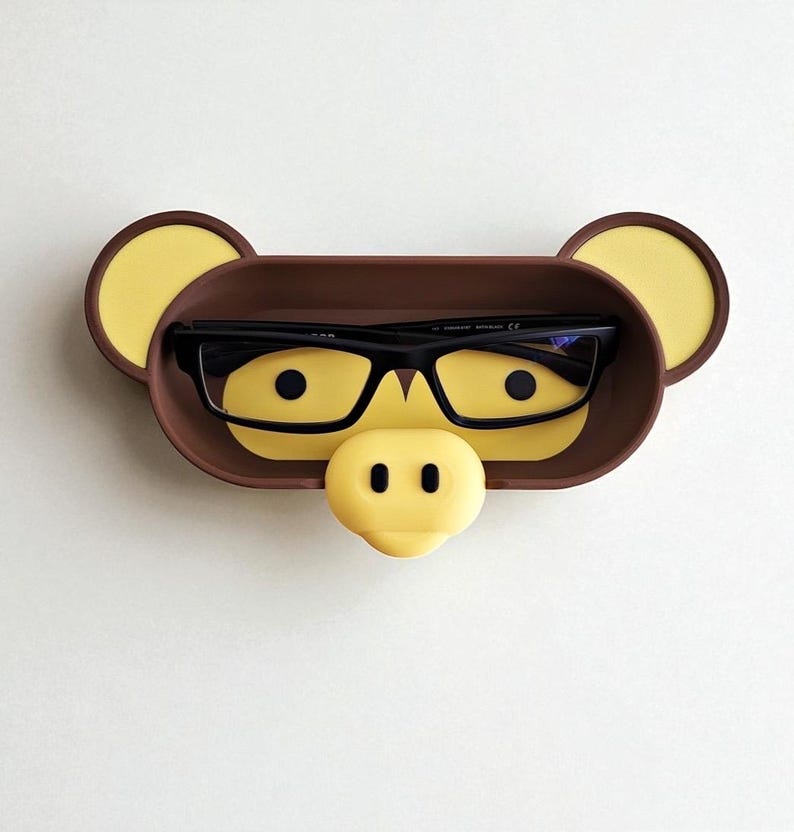 May include: A brown and yellow eyeglasses holder shaped like a monkey face. The holder has a tray for glasses, yellow ears, and a yellow nose. Black eyeglasses rest inside the tray.