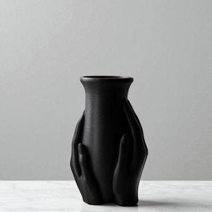 Op de afbeelding: Een matzwarte vaas met een getextureerd oppervlak. De vaas heeft een uniek ontwerp met twee handen die rond de body zijn gebeeldhouwd, wat een opvallend visueel effect cre&euml;ert. Een decoratief item.