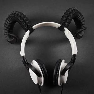 May include: White and black headphones with black, textured ram horns attached to the headband. The headphones have a white headband and earcups, with black ear pads. The background is a solid dark grey.