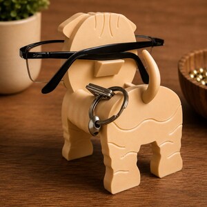 May include: A tan-colored dog-shaped eyeglasses and key holder. The holder has a slot for glasses and a ring for keys. Black-framed eyeglasses rest on the holder. The holder is made of a solid material and has a textured surface.