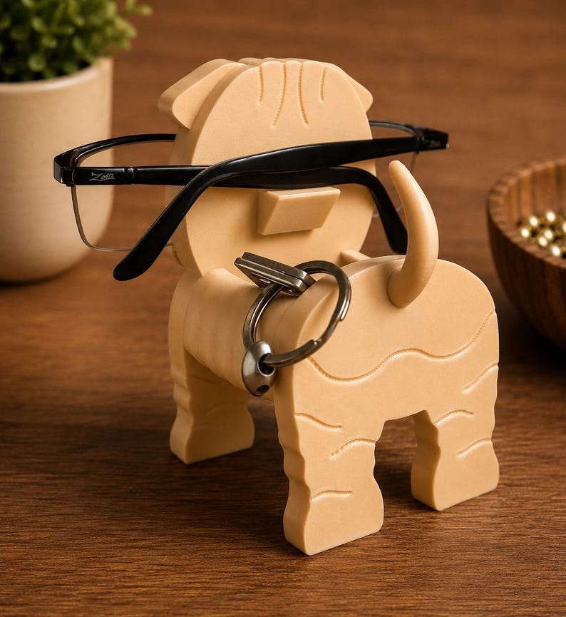 May include: A tan-colored dog-shaped eyeglasses and key holder. The holder has a slot for glasses and a ring for keys. Black-framed eyeglasses rest on the holder. The holder is made of a solid material and has a textured surface.