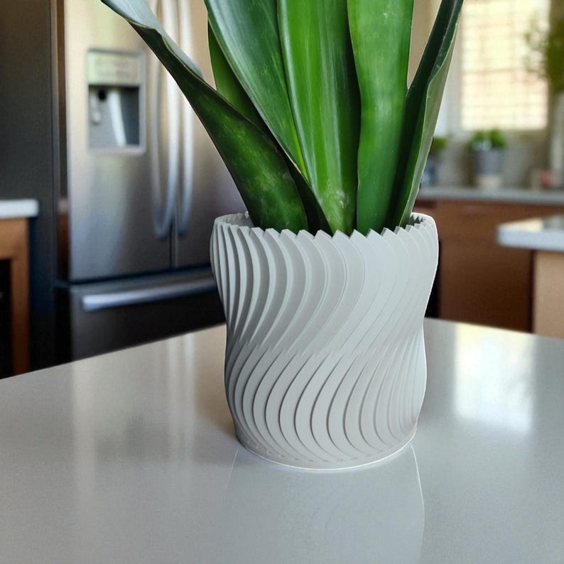 May include: A white, spiral-patterned planter pot holding a green plant with long, upright leaves. The pot has a textured, wavy design and sits on a white countertop. The background includes a stainless steel refrigerator and kitchen cabinets.