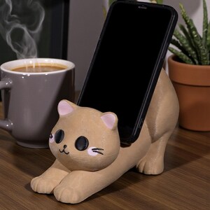 May include: A tan cat-shaped phone holder with a black phone in place. The holder has pink accents on the ears and cheeks, with black eyes and whiskers. The phone is held at an angle, and the holder is on a wooden surface.