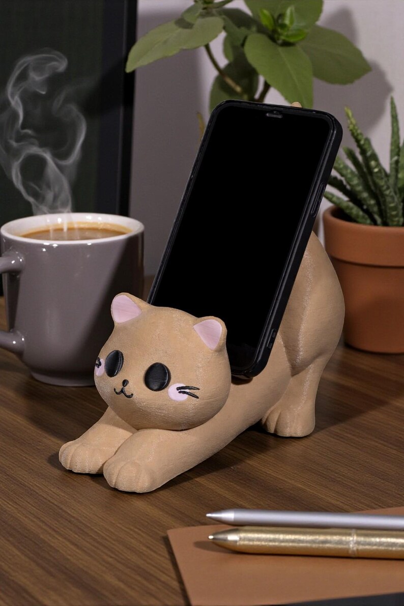 May include: A tan cat-shaped phone holder with a black phone in place. The holder has pink accents on the ears and cheeks, with black eyes and whiskers. The phone is held at an angle, and the holder is on a wooden surface.