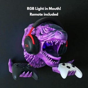 May include: A purple dinosaur head with RGB lighting in its mouth, holding two game controllers. A black and red headset rests on the head. The text "RGB Light in Mouth! Remote included" is at the top.