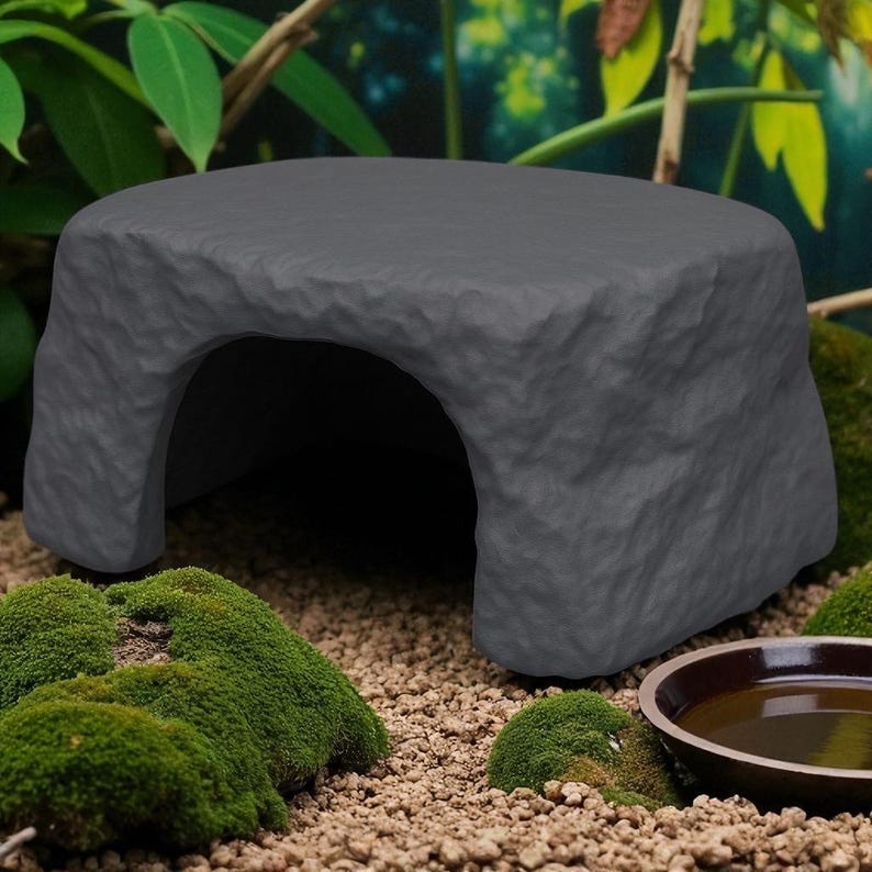 May include: A gray, rock-like reptile hideout with an arched opening. The hideout is rectangular and sits on a bed of small pebbles and green moss. A small, brown bowl is visible in the foreground.