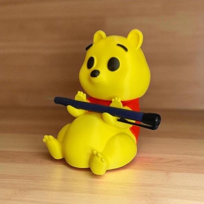 May include: A yellow Winnie the Pooh figurine holding a blue pen. The figurine has a red shirt and black eyes. The background is a wooden surface.
