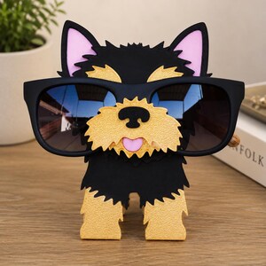 May include: A black and tan dog-shaped eyeglass holder with a pink tongue and ears. The holder is wearing black sunglasses. The dog is standing on a wooden surface.