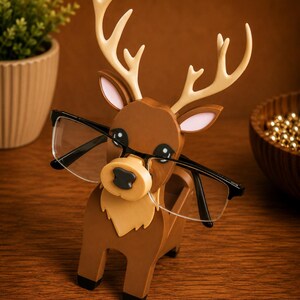 May include: A wooden reindeer-shaped glasses holder in shades of brown, tan, and cream. The holder has a slot to hold glasses, with the frames resting on the reindeer's nose. The reindeer has large black eyes and cream-colored antlers. The glasses are black-framed.