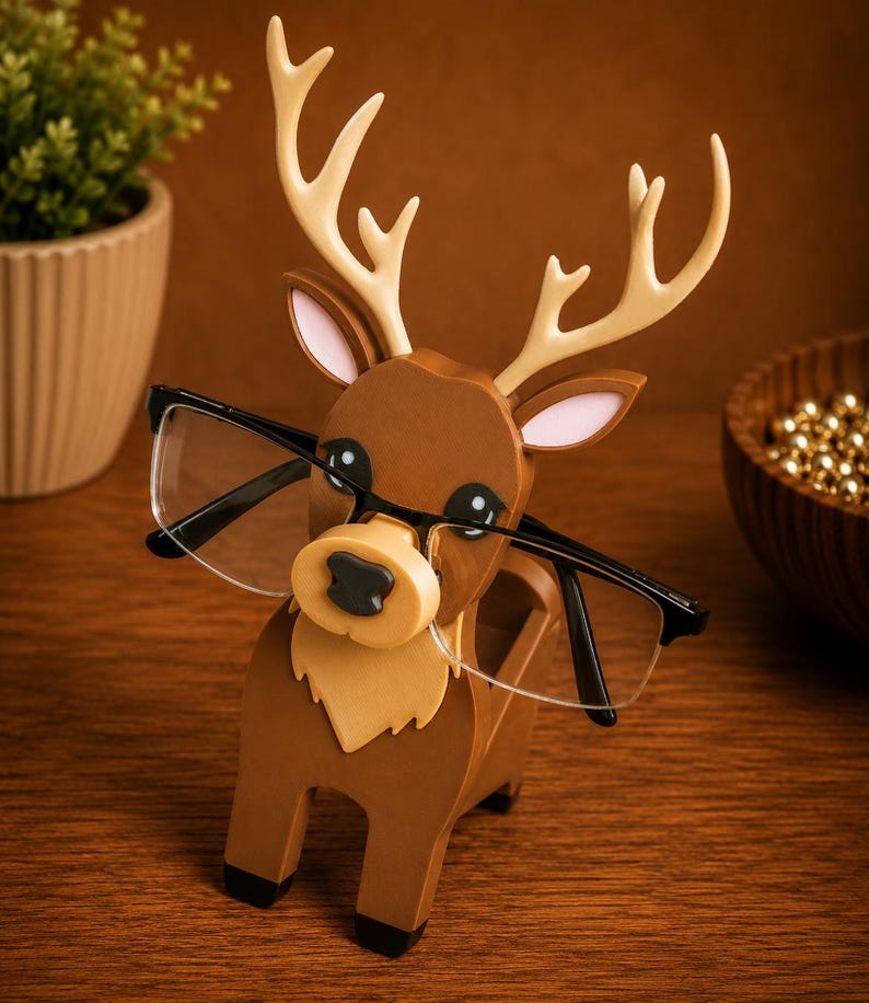 May include: A wooden reindeer-shaped glasses holder in shades of brown, tan, and cream. The holder has a slot to hold glasses, with the frames resting on the reindeer's nose. The reindeer has large black eyes and cream-colored antlers. The glasses are black-framed.
