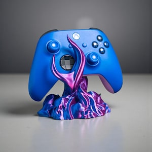 May include: A blue Xbox controller rests on a decorative stand. The controller has a black D-pad and buttons. The stand is a swirling design of purple and blue. The stand is designed to hold the controller.