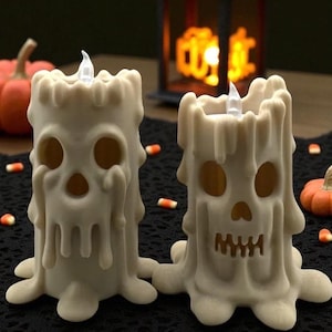 May include: Two off-white Halloween candles with skull faces and dripping wax details. The candles have a flickering LED light at the top. Candy corn and small pumpkins are scattered around the candles on a black surface.