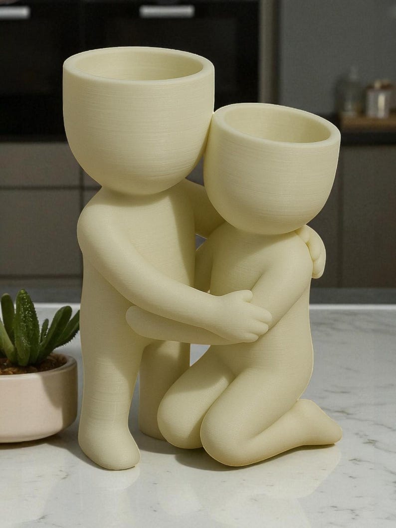 K&ouml;nnte beinhalten: Eine cremefarbene Keramikskulptur, die zwei sich umarmende Figuren darstellt. Eine Figur steht, die andere kniet, beide mit offenen K&ouml;pfen, die als Pflanzgef&auml;&szlig;e konzipiert sind. Im Hintergrund ist eine kleine Sukkulentenpflanze in einem rosa Topf zu sehen.