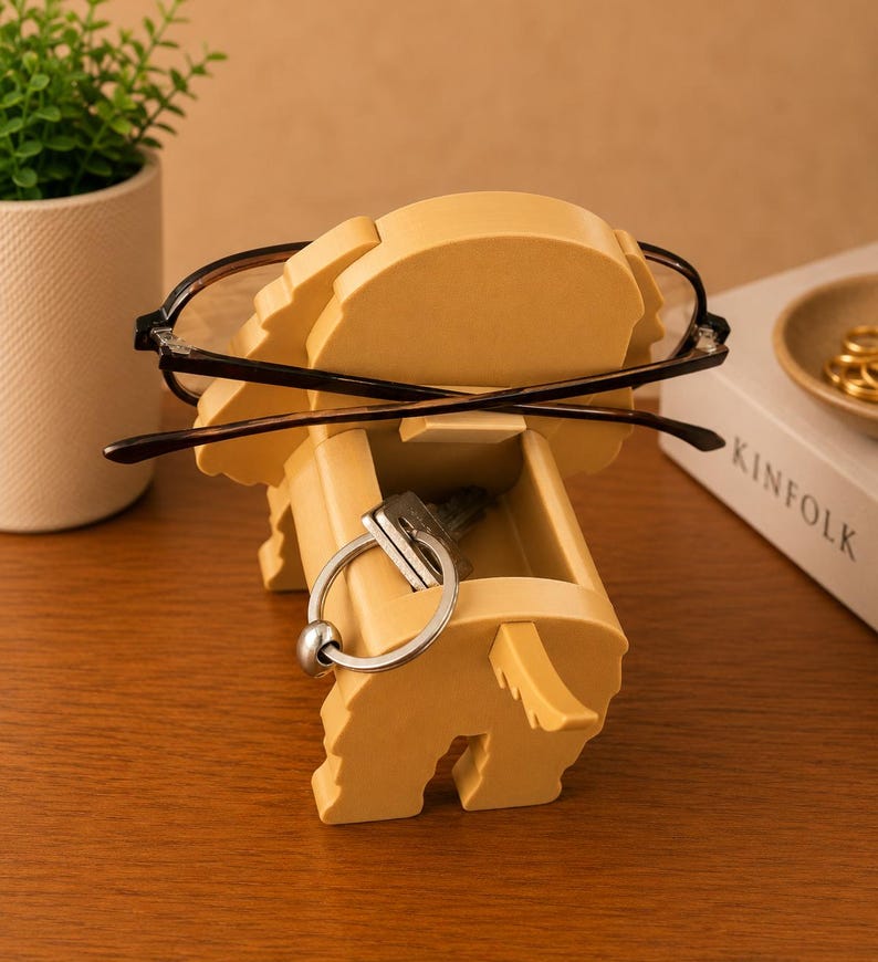 K&ouml;nnte beinhalten: Ein hellgelber, elefantenf&ouml;rmiger Organizer, der Brillen und Schl&uuml;ssel h&auml;lt. Der Elefant hat eine kleine Schublade zur Aufbewahrung. Die Brille hat einen schwarzen Rahmen. Eine kleine Topfpflanze und ein Buch befinden sich im Hintergrund.