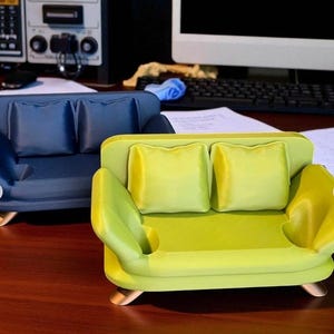 May include: Two miniature sofas, one navy blue and one lime green, each with two square pillows. The green sofa has a small circular indentation on the arm. Both sofas have gold-colored feet.