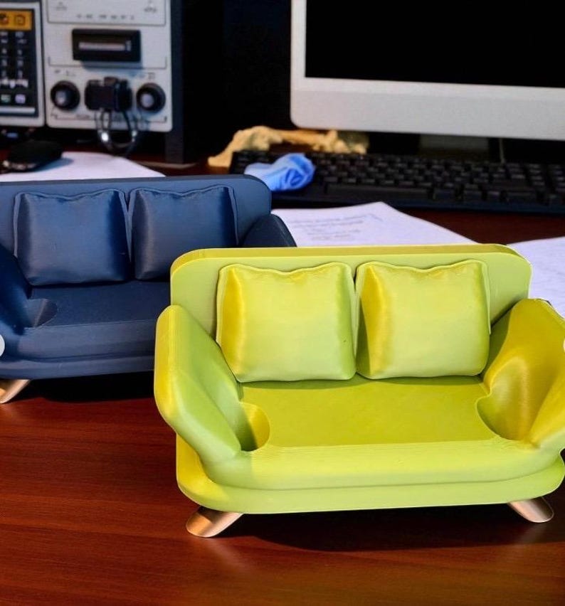May include: Two miniature sofas, one navy blue and one lime green, each with two square pillows. The green sofa has a small circular indentation on the arm. Both sofas have gold-colored feet.