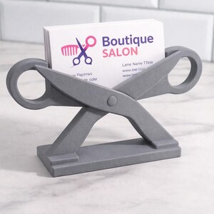 May include: A gray scissor-shaped business card holder. The holder has a base and two large scissor blades with circular handles. White business cards with the text "Boutique Salon" are in the holder.